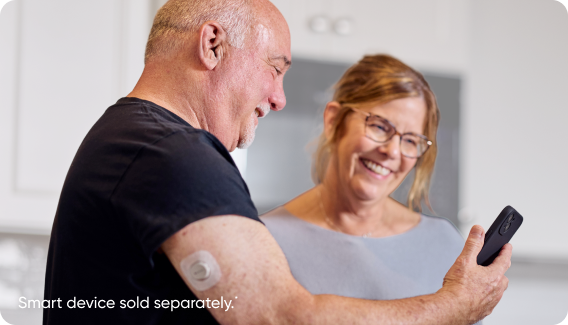 Dexcom G7 user showing his phone to another person. Includes caption that says "Smart devices sold separately."